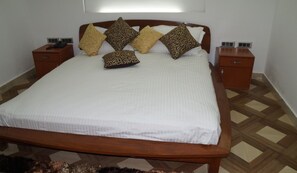 Una cama bien hecha con una colcha blanca y almohadas estampadas, flanqueada por mesitas de noche de madera con un teléfono y un cajón pequeño.