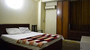A single bed with a patterned blanket, a wooden headboard, an air conditioning unit, and a wooden wardrobe. - Shamrock Hotel (Lahore)