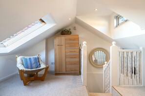 Une pièce avec un plafond en pente, une commode en bois avec des tiroirs, une chaise en osier avec un coussin, un miroir rond et un escalier blanc avec une balustrade.