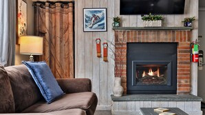 A living room with a brown sofa, a blue pillow, a fireplace, a mounted TV, and a fire extinguisher. - The Hideaway Retreat at Mountain Green 1 BR/1 BA Condo, Steps to the Mountain, Amenities. (Killington)
