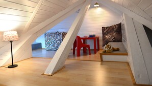An attic room with a sloped ceiling, a small table with a chair, a sofa with a tiger plush toy, and a lamp.