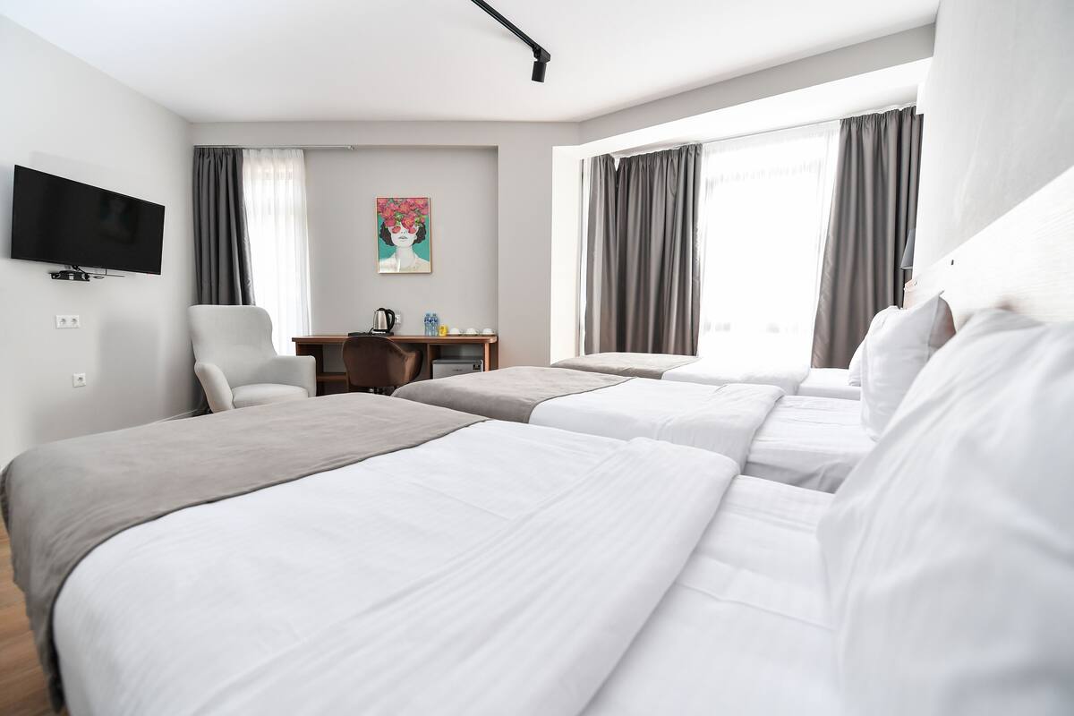 A hotel room with three beds, a desk, a chair, a flat-screen TV, and a painting on the wall.