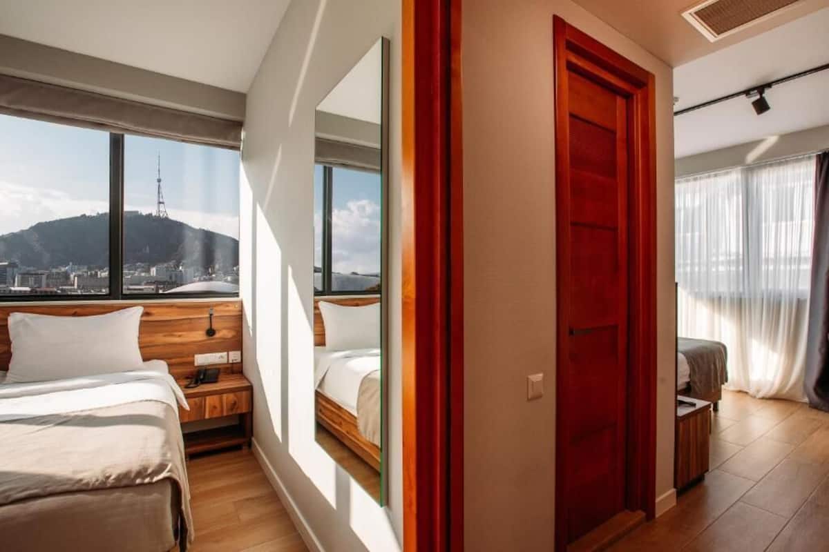 A hotel room with a bed, a nightstand, a window with a city view, and a red door.
