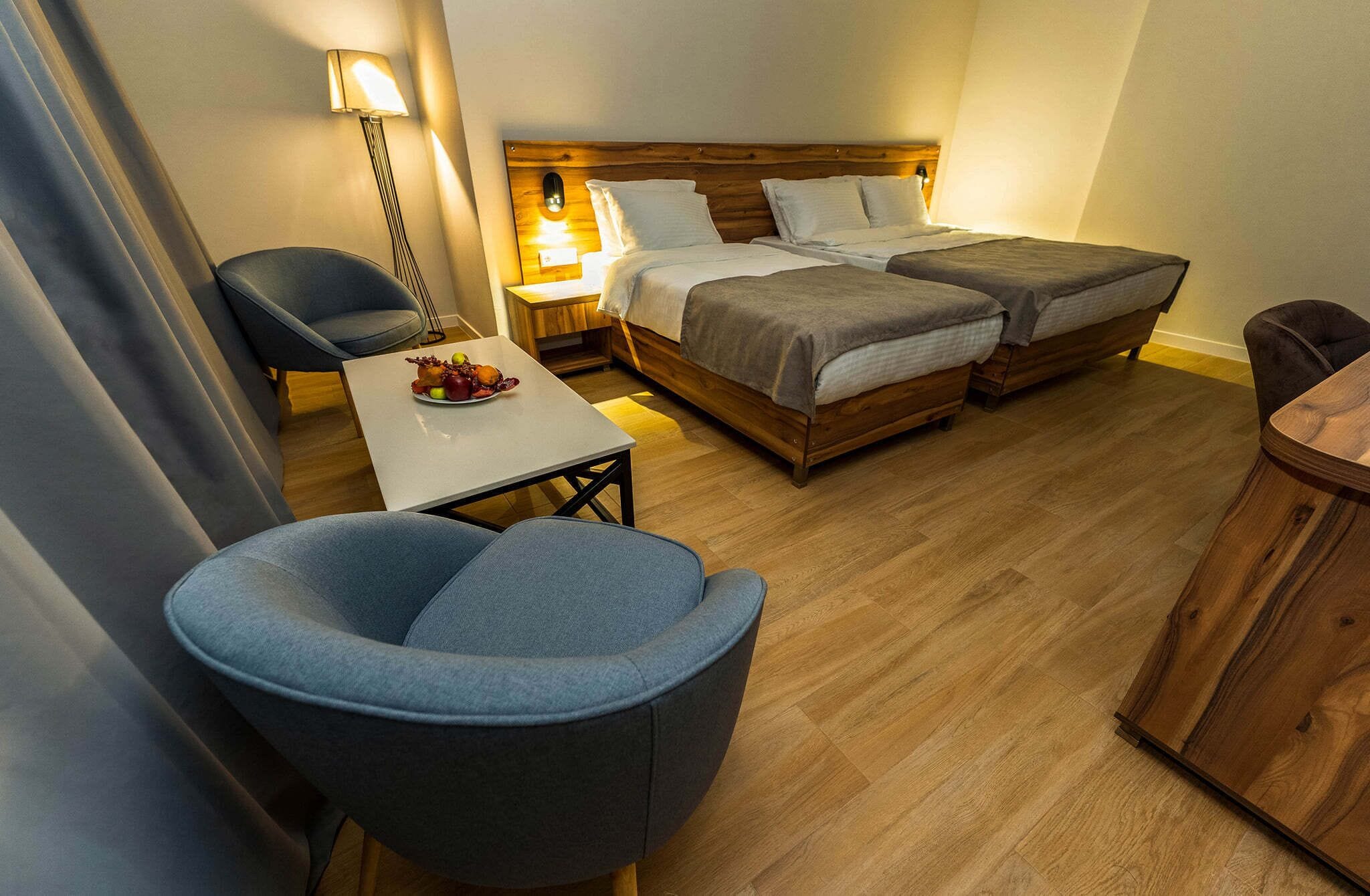 A hotel room with two beds, a desk, a chair, and a small table with fruit.