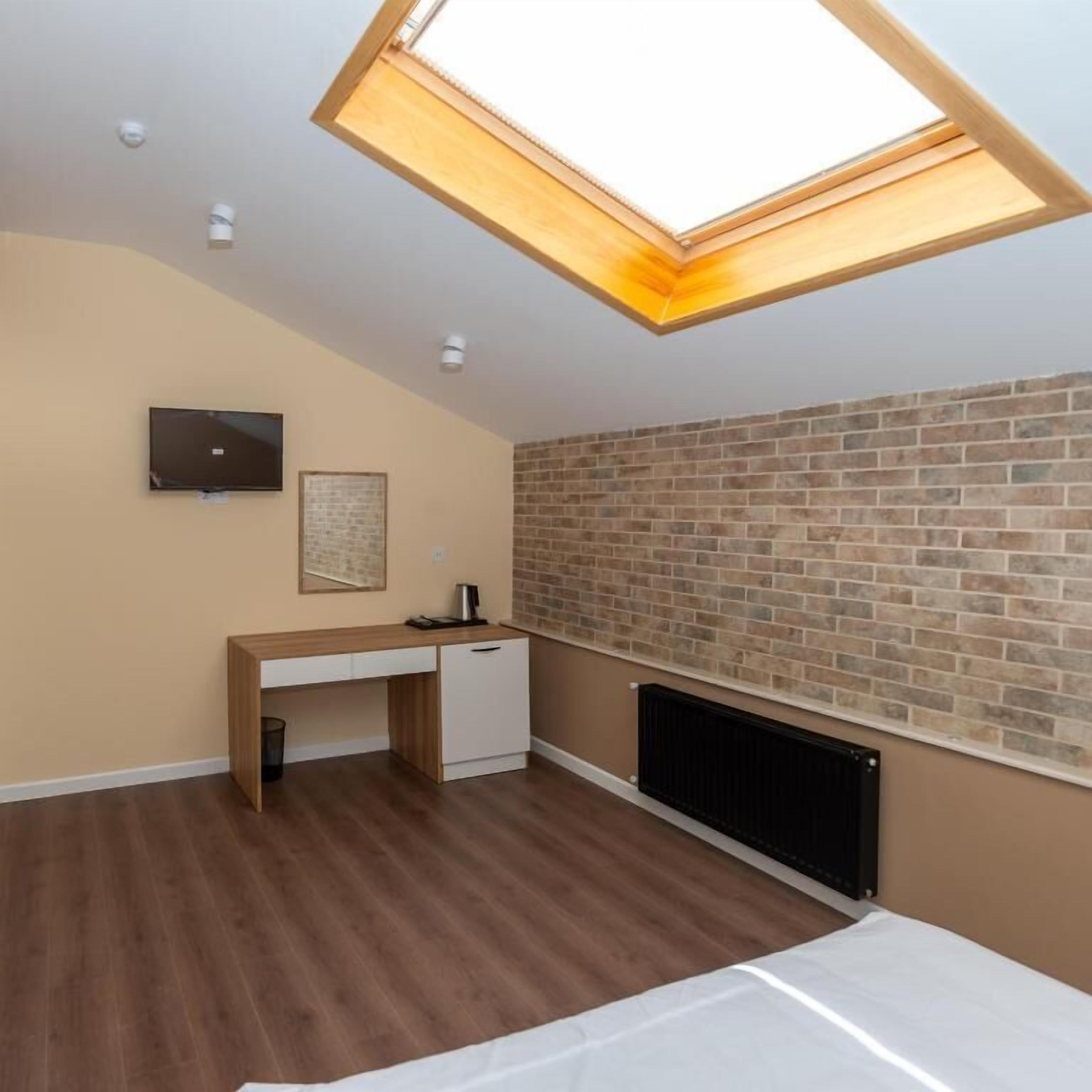A bedroom with a skylight, a brick wall, a desk, a radiator, and a television.