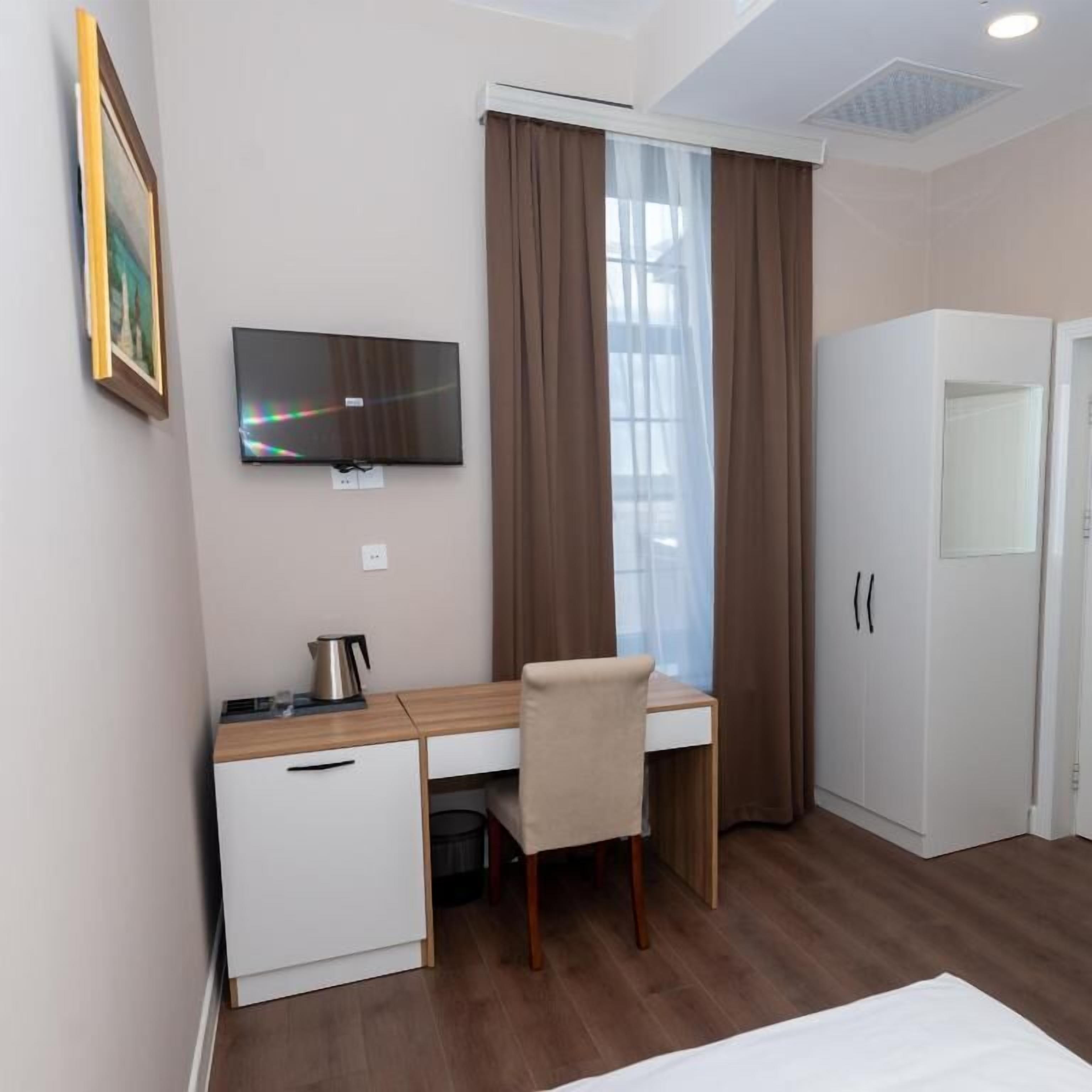 A hotel room with a wooden desk, a chair, a television mounted on the wall, a wardrobe, and a window with curtains.