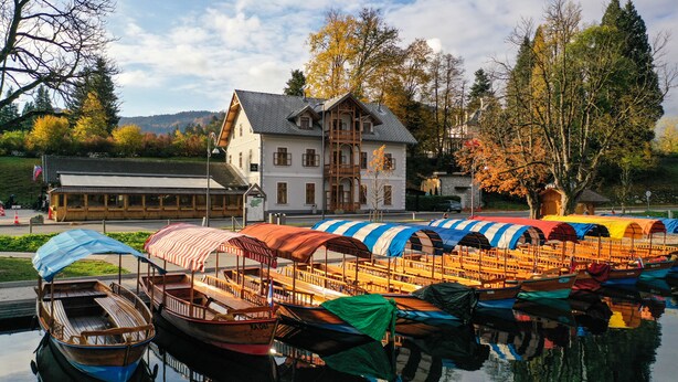 A lakeside hotel with boats docked in front, a large building with a balcony, and autumn trees in the background. - HOTEL STARKL HERITAGE & UNIQUE (Bled)