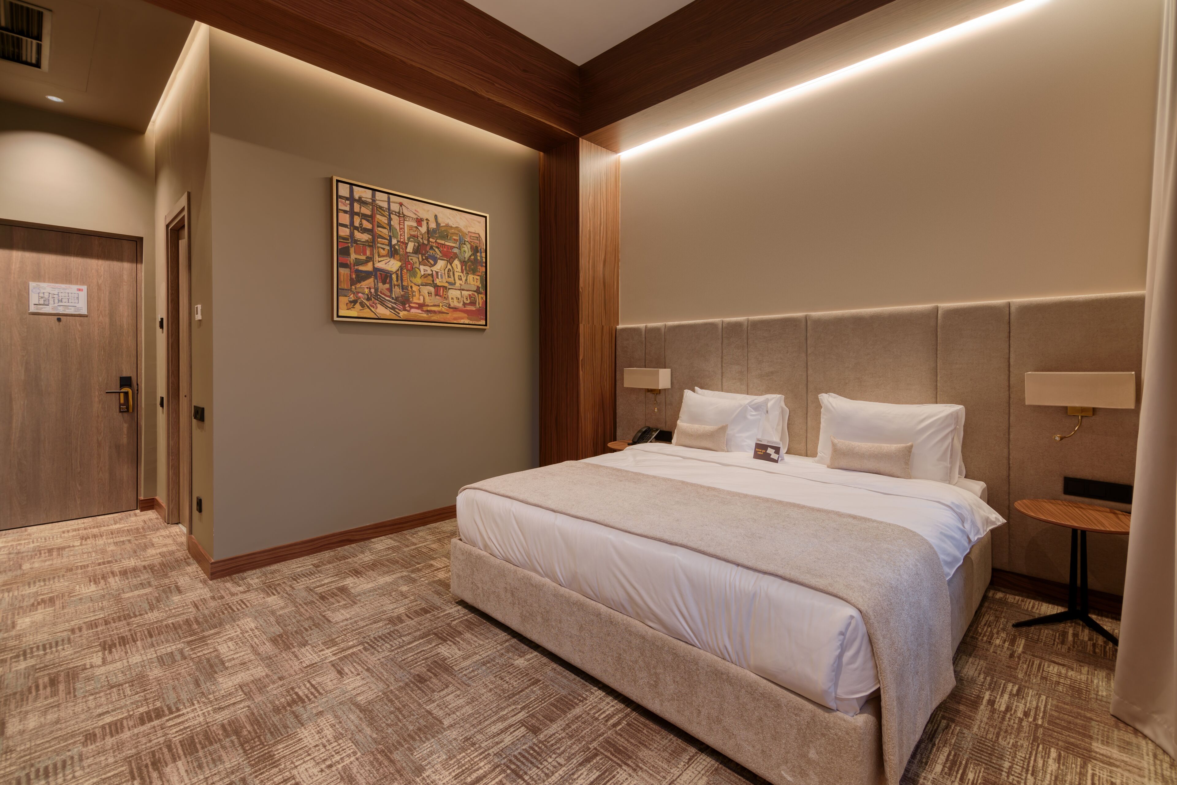 A hotel room with a large bed, a headboard, a bedside table, a wall-mounted lamp, and a painting on the wall.
