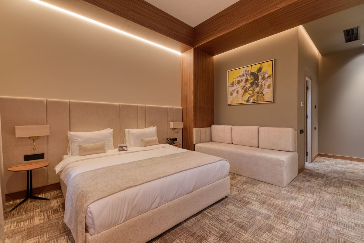A modern hotel room with a large bed, a sofa, a small table, and a painting on the wall.