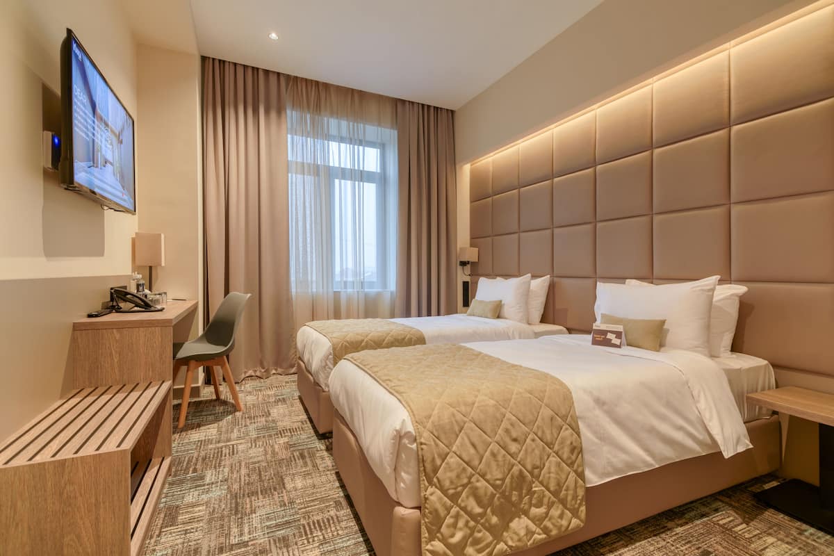 A hotel room with two beds, a desk with a chair, a TV, and a large window with curtains.