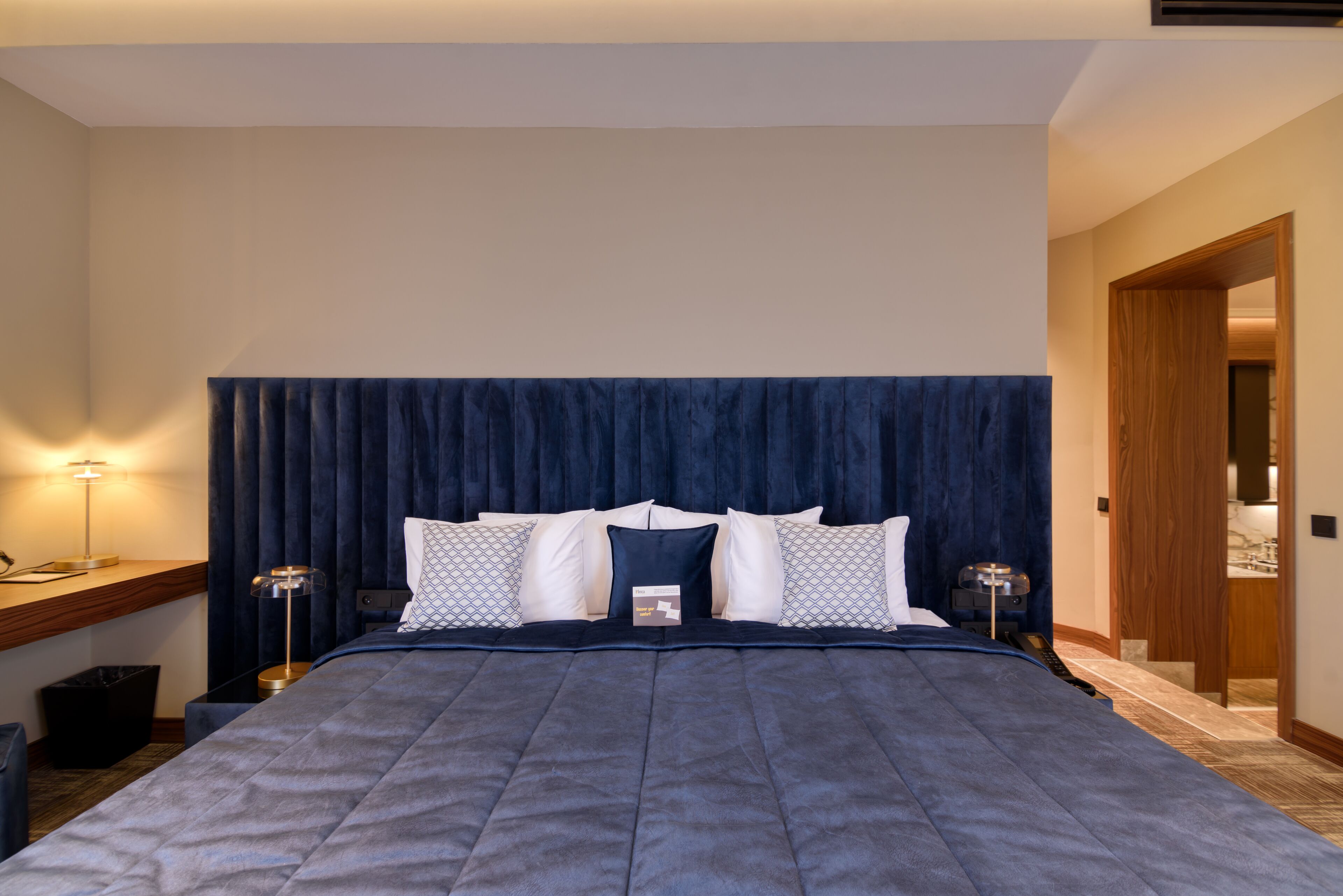 A hotel room with a large bed, a wooden desk, a lamp, and a door leading to another room.