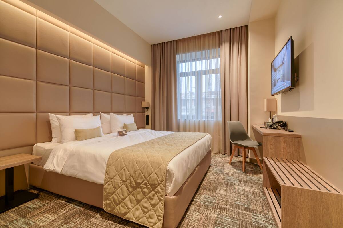 A modern hotel room with a large bed, a desk, a chair, and a TV mounted on the wall.