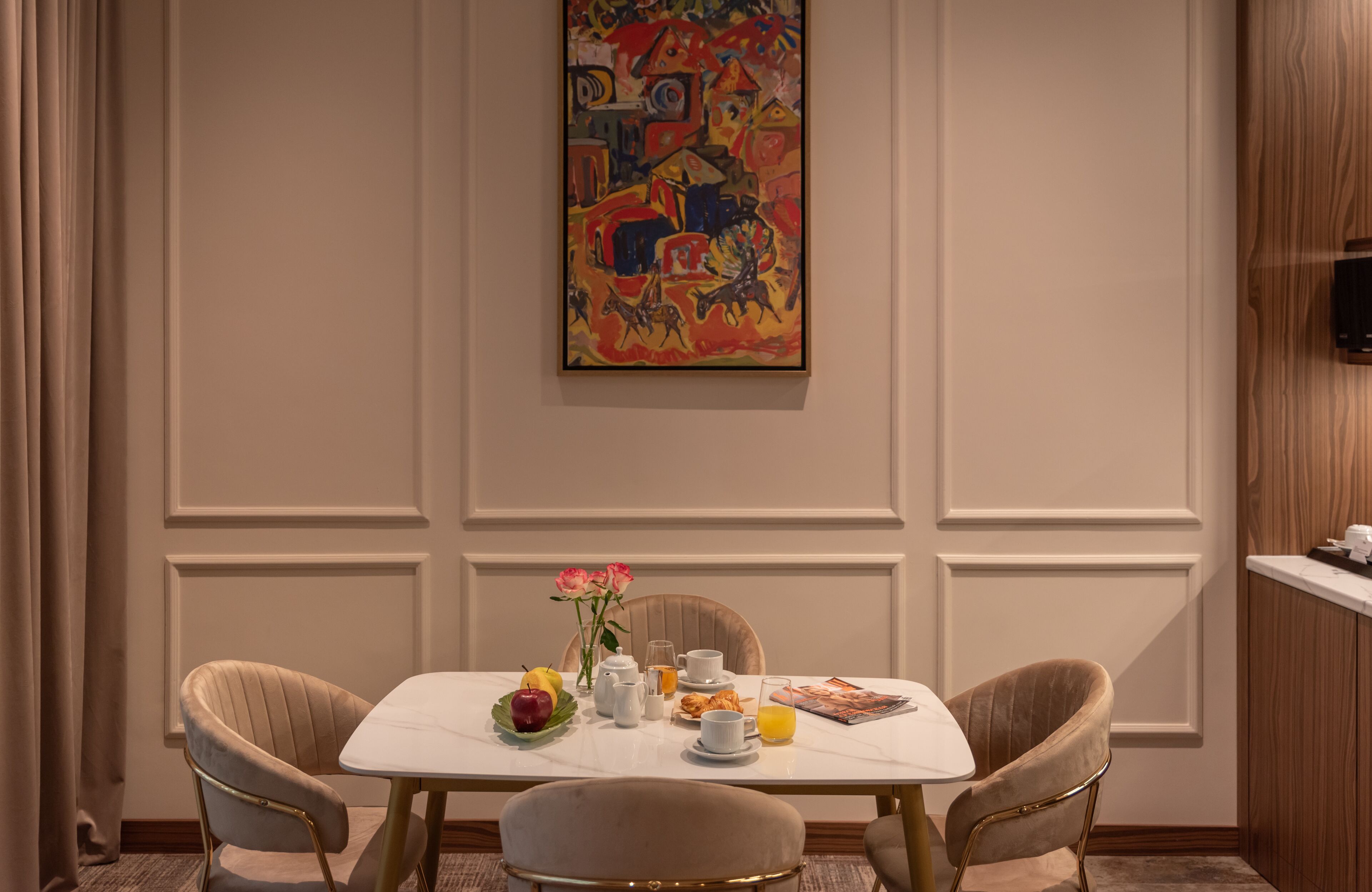 Un comedor con una mesa de mármol, dos sillas tapizadas, un jarrón con flores, un frutero, una vajilla para café y un cuadro abstracto de colores en la pared.