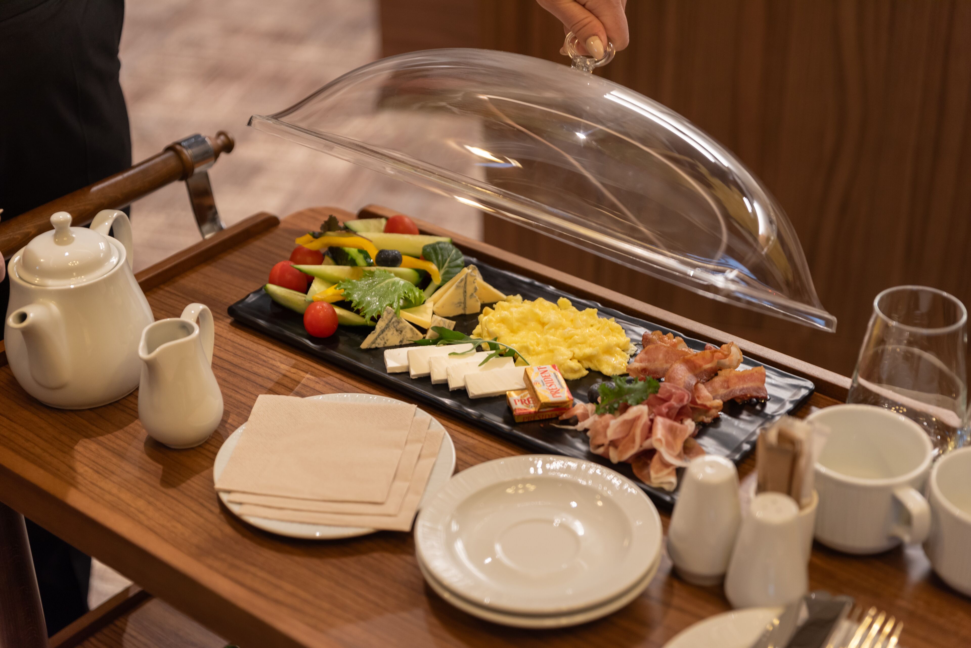 A breakfast buffet with a variety of foods, including eggs, bacon, and cheese, served on a wooden table with plates, cups, and a pitcher.