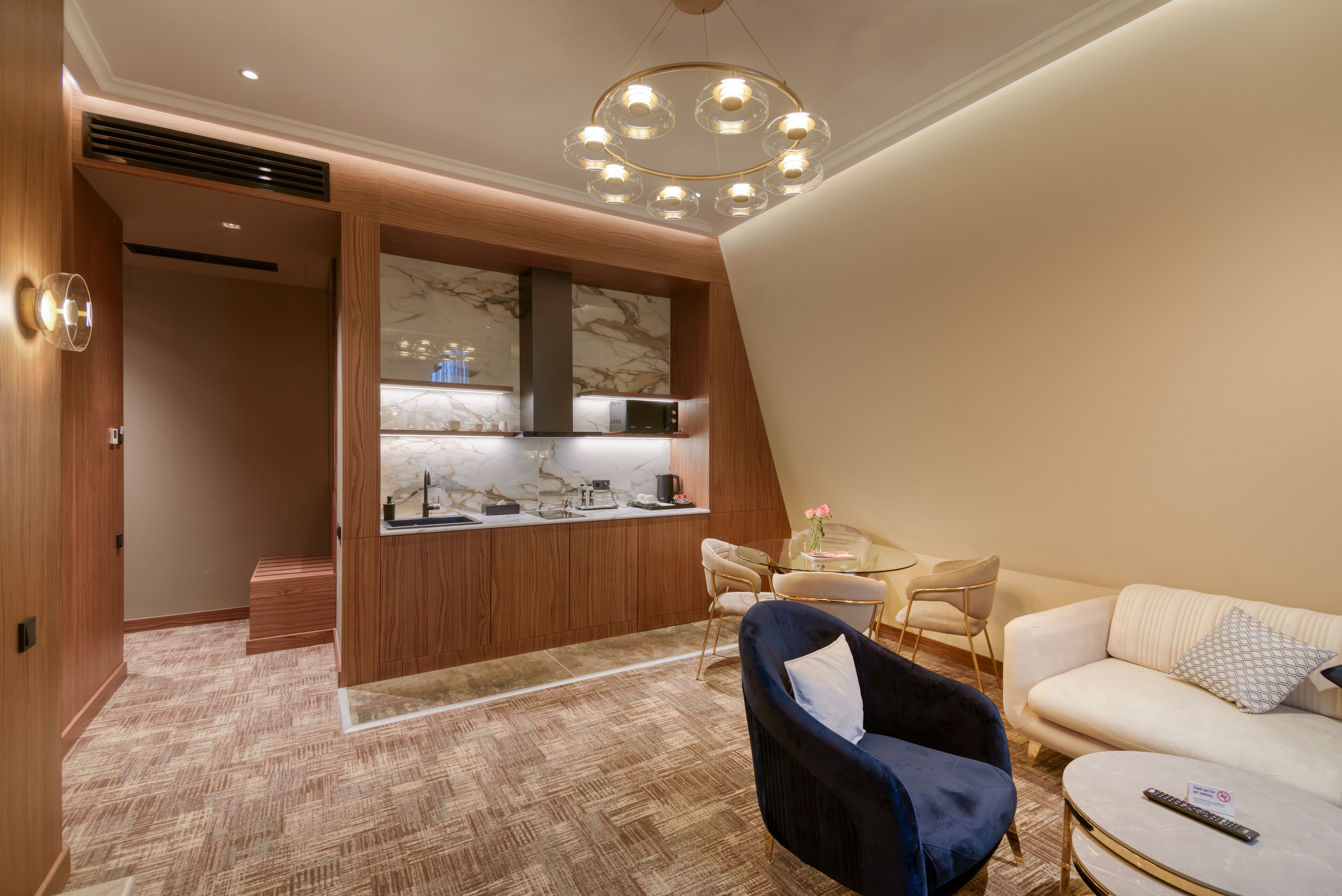 A modern hotel room with a kitchen, a dining area, and a living space with a sofa and armchair.