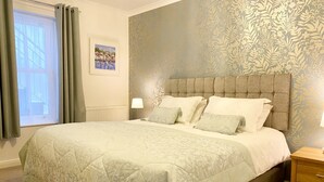 A neatly made bed with a patterned bedspread, a headboard with a floral wallpaper design, and a window with curtains. - Spacious, one bedroom, pet friendly apartment with large terrace overlooking the town. (Torquay)