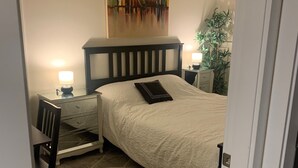 Camera da letto con un letto, un comodino, una sedia, una lampada, un quadro appeso e una pianta.