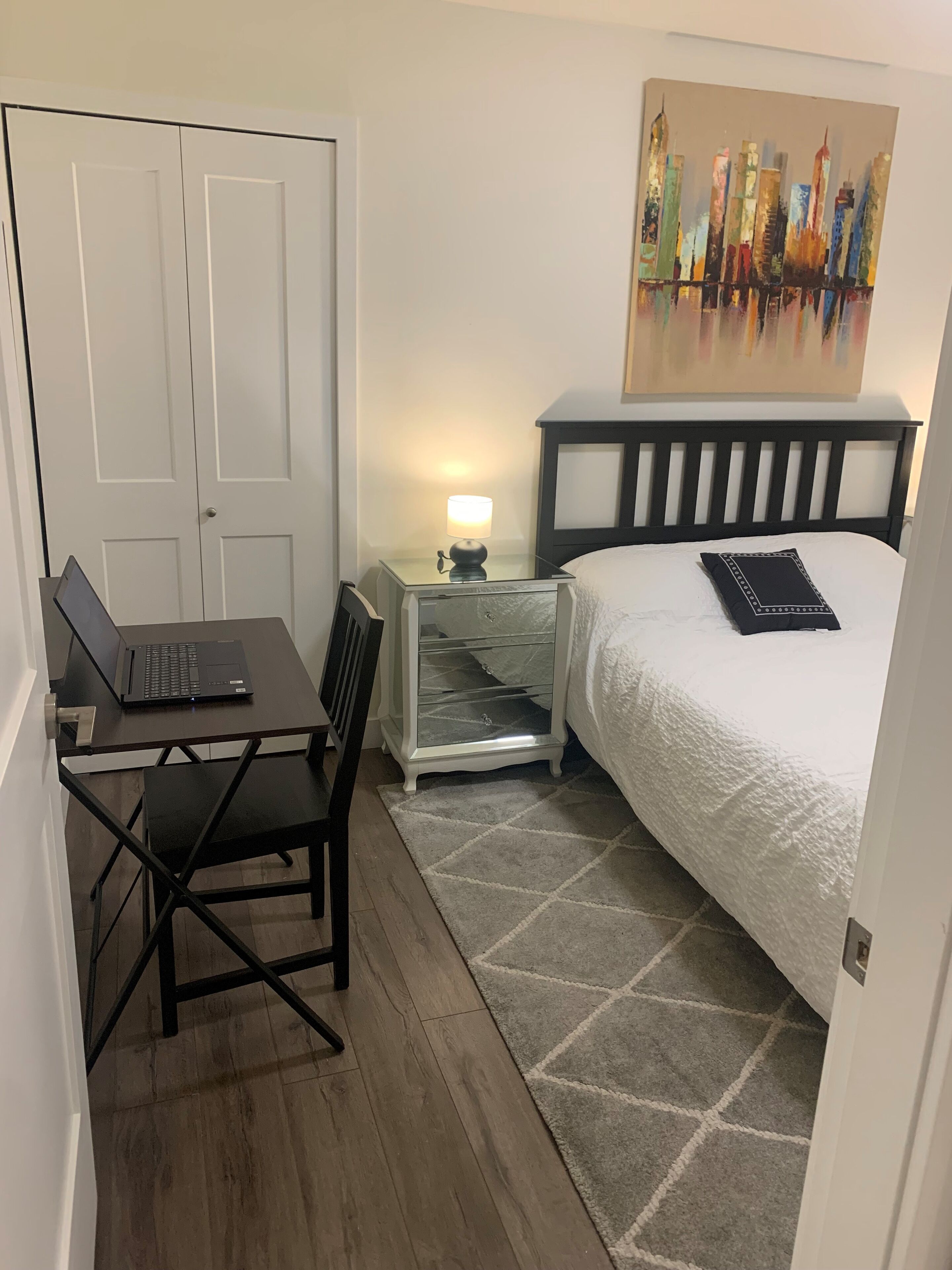 Habitación de hotel con una cama, un escritorio con una computadora portátil, una mesita de noche con una lámpara y un cuadro en la pared.