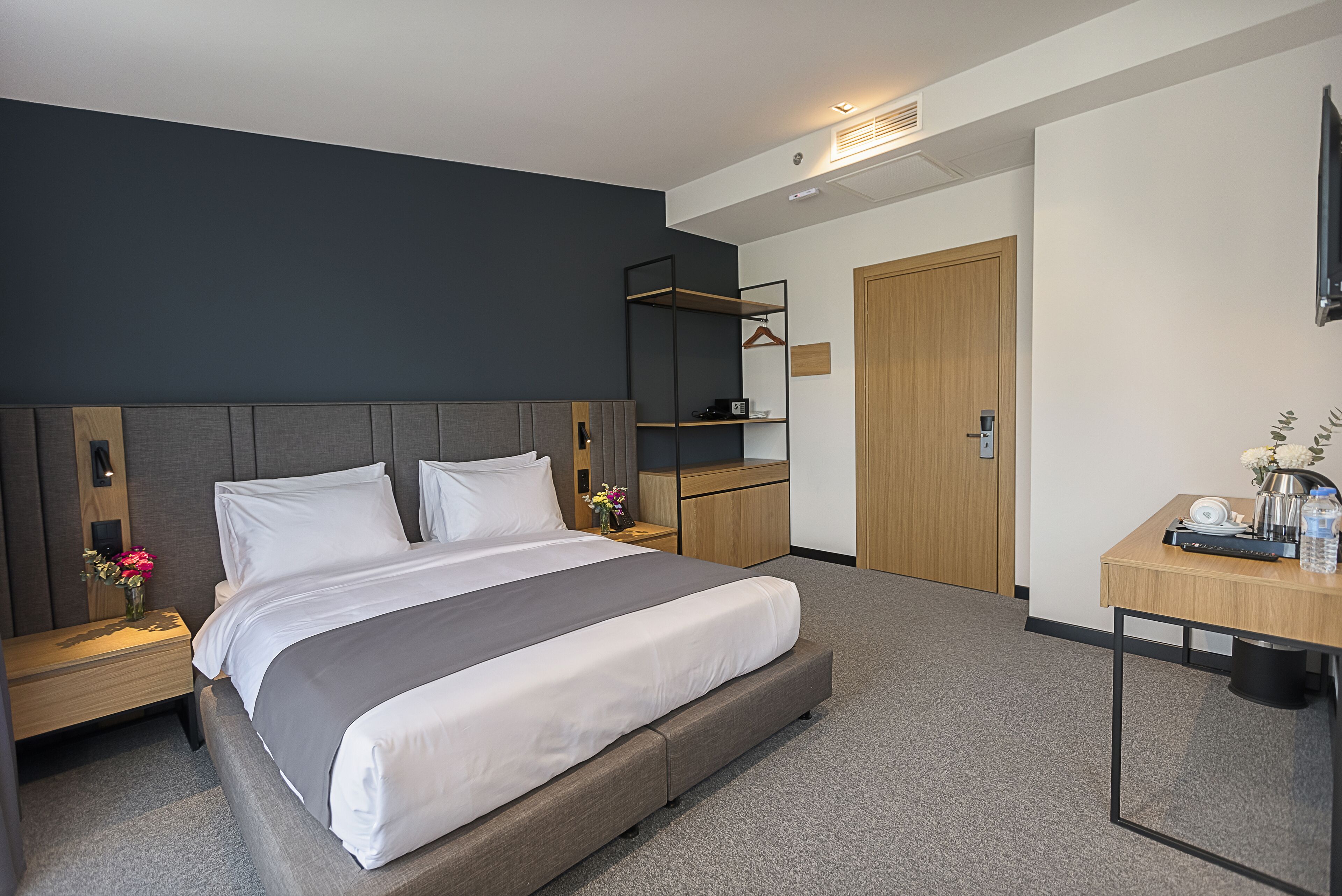 A modern hotel room with a large bed, bedside tables, a wardrobe, and a desk with a chair.