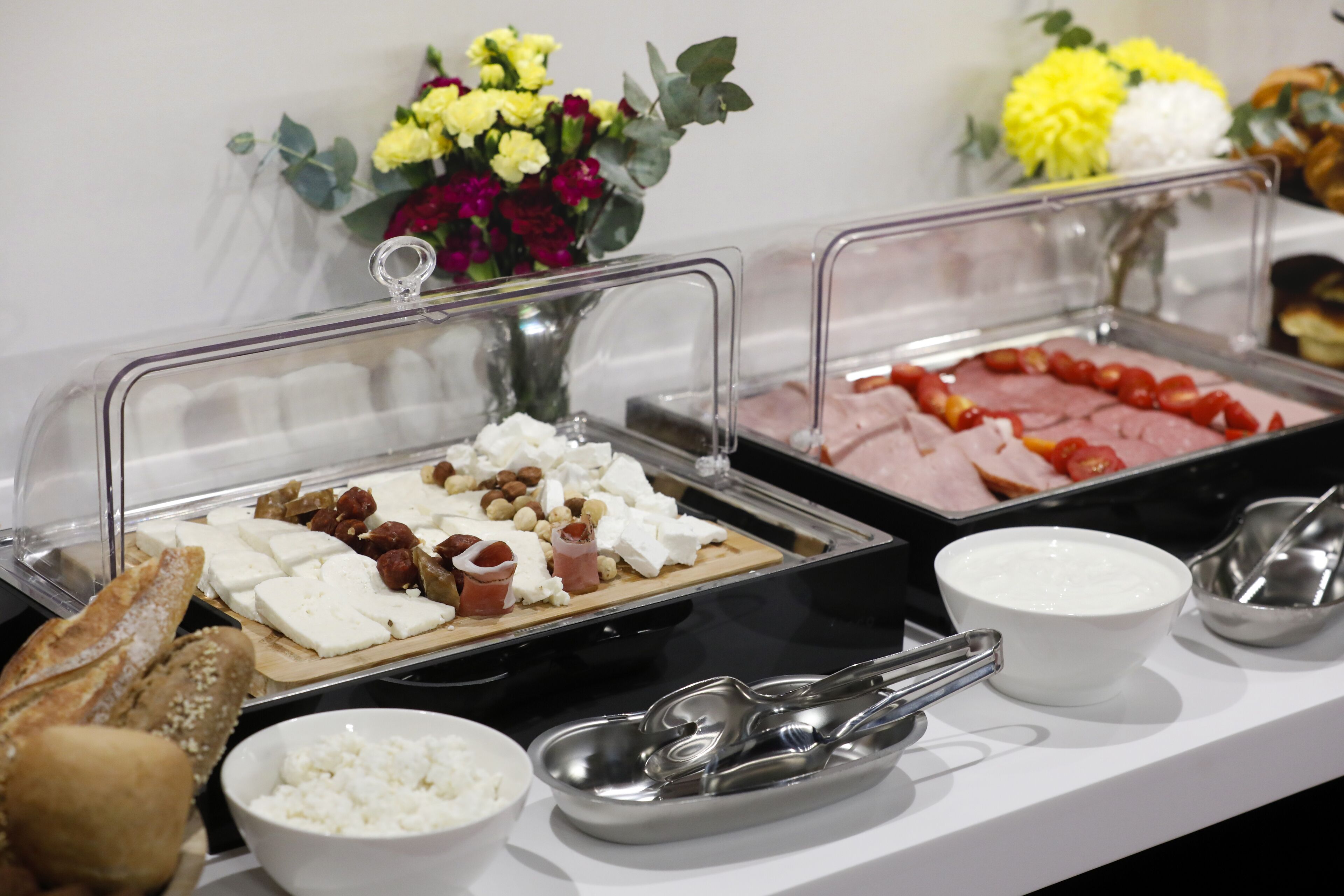 Een buffet met diverse soorten eten, waaronder kazen, vleeswaren en brood, gepresenteerd op een toonbank, met een bloevas op de achtergrond.