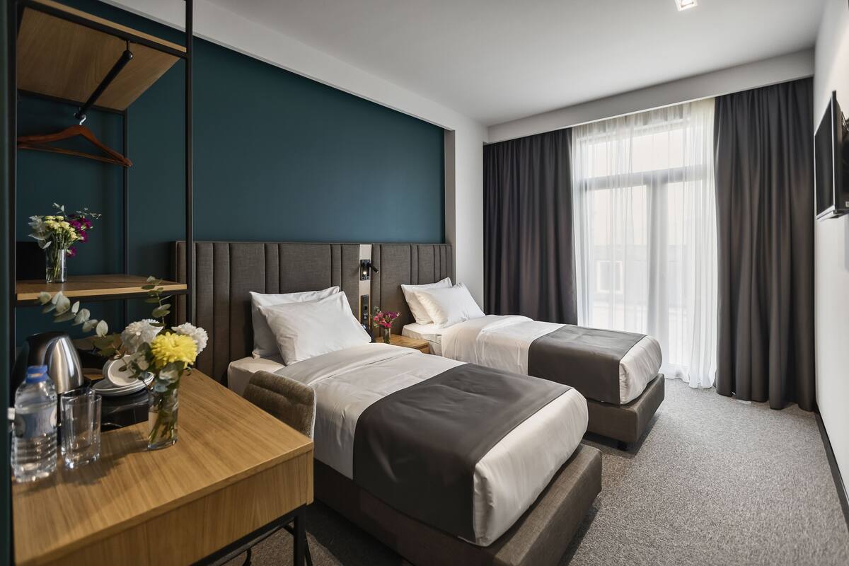 A hotel room with two beds, a desk with a chair, a mirror, and a window with curtains.