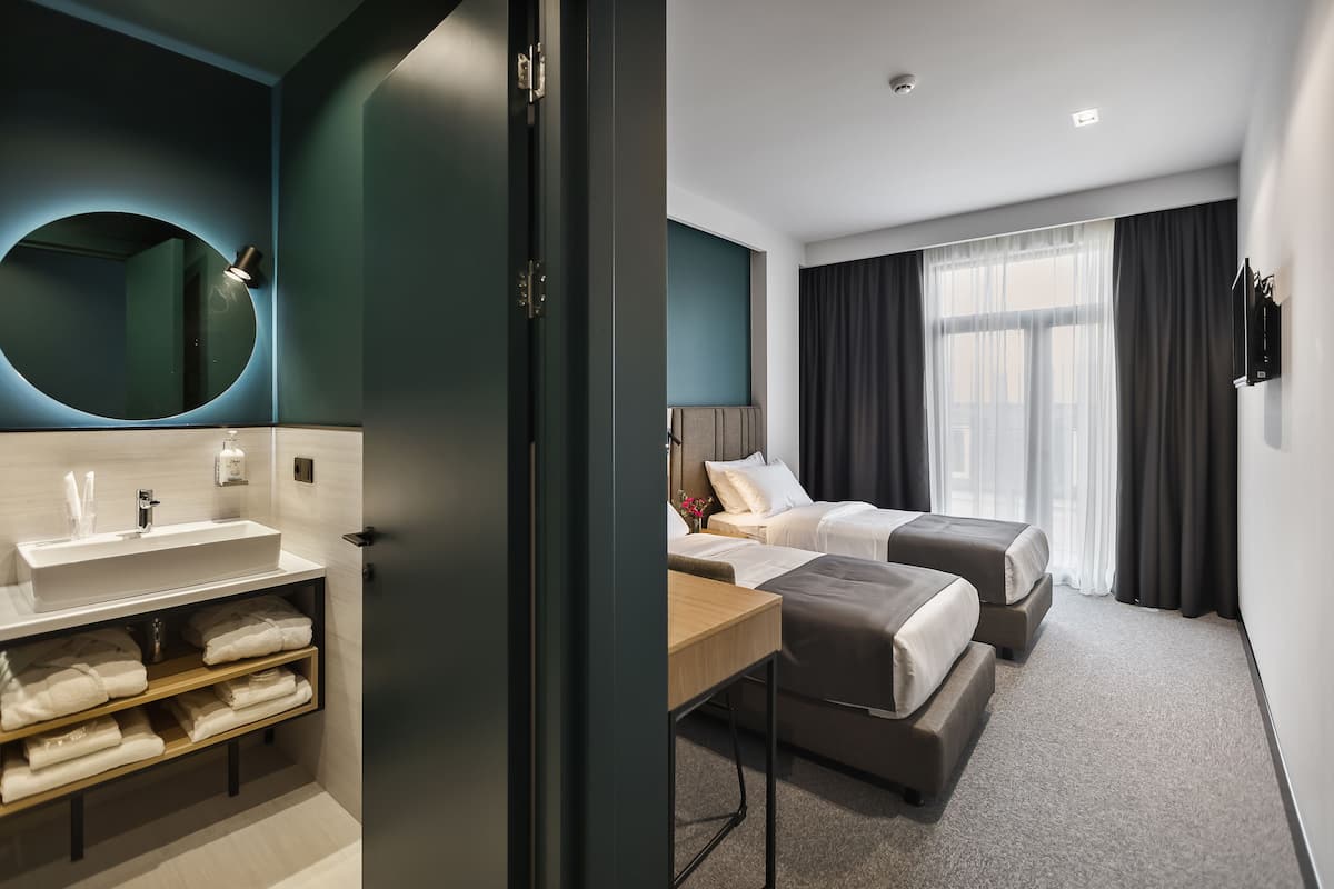 A modern hotel room with a large round mirror, a white sink, a wooden shelf with towels, a desk, two beds, and a large window with curtains.
