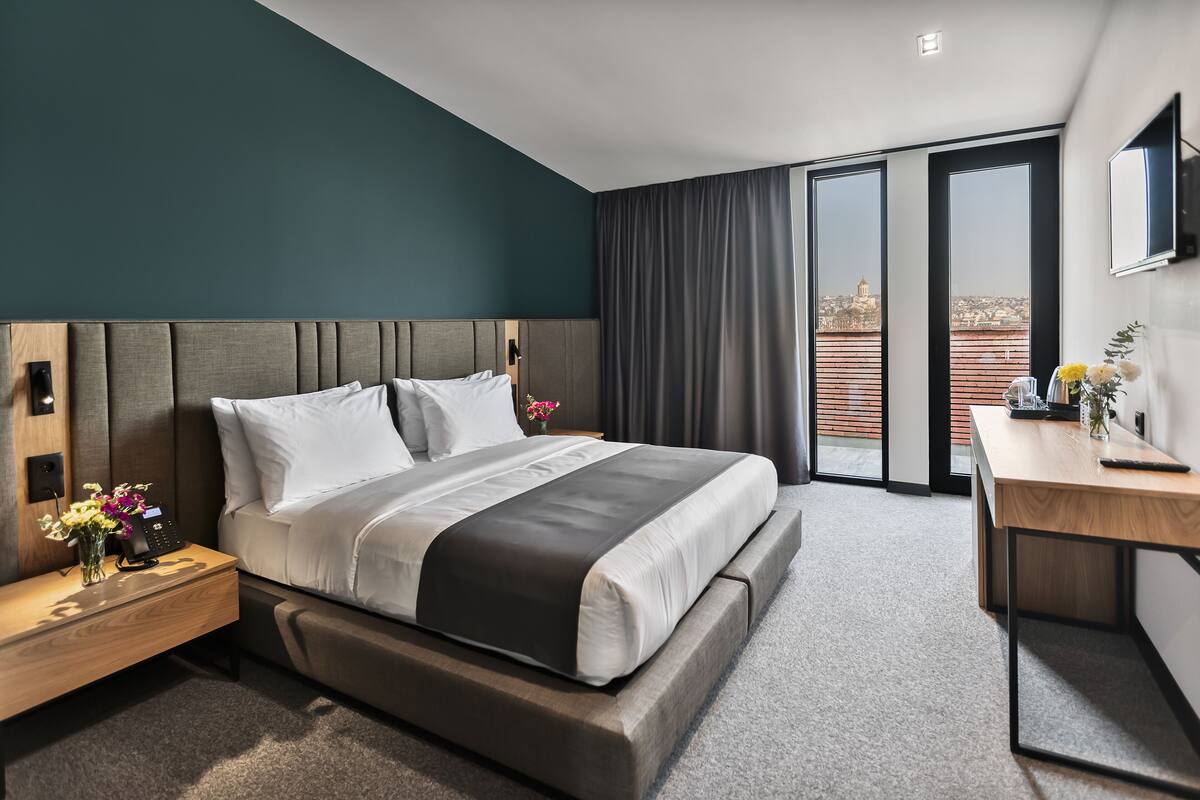 A modern hotel room with a large bed, a desk, and a view of the city through the window.