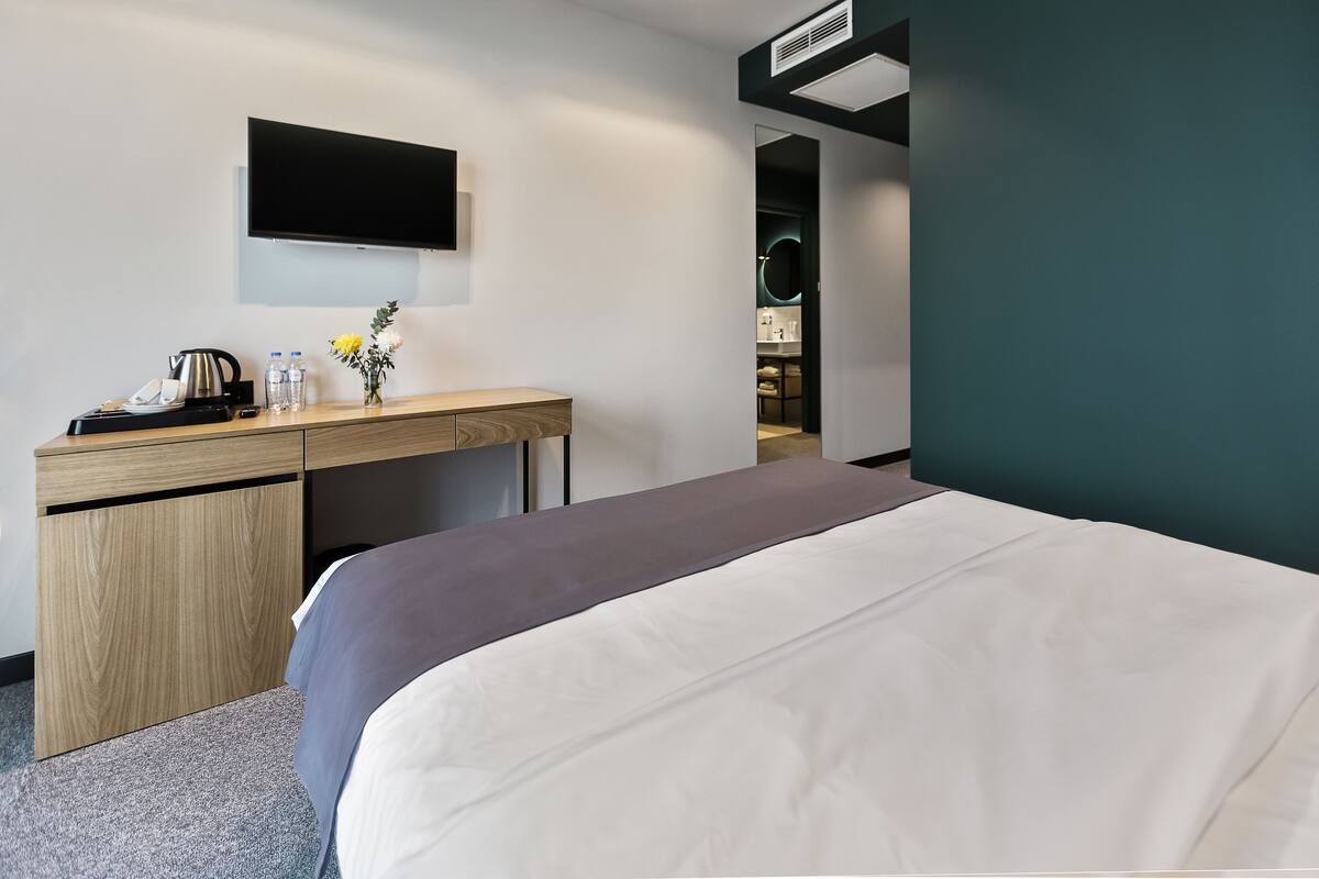 A hotel room with a bed, a desk with a flat-screen TV, a coffee maker, and a vase with flowers.