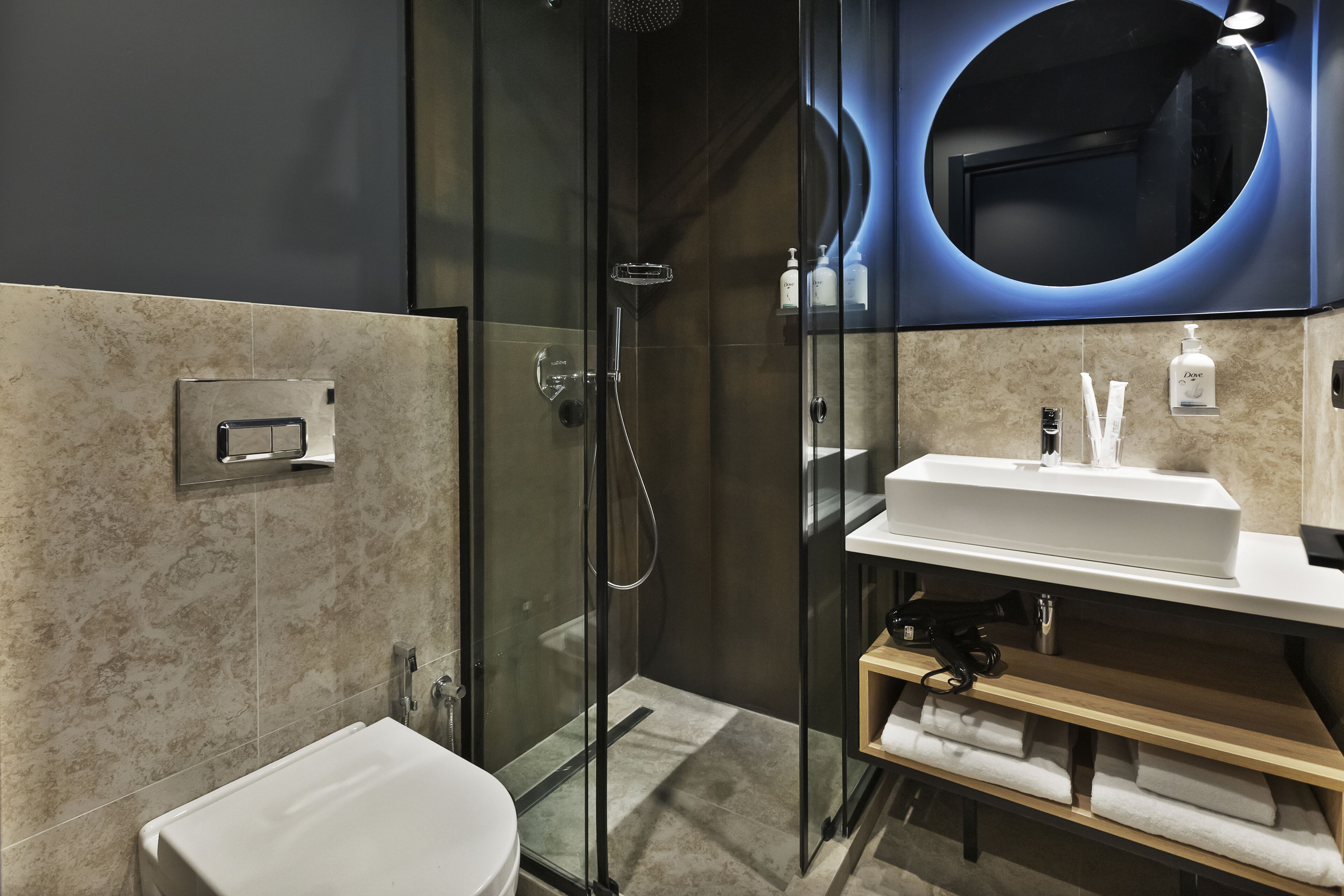 A modern bathroom with a glass-enclosed shower, a white sink, and a mirror with a circular light.