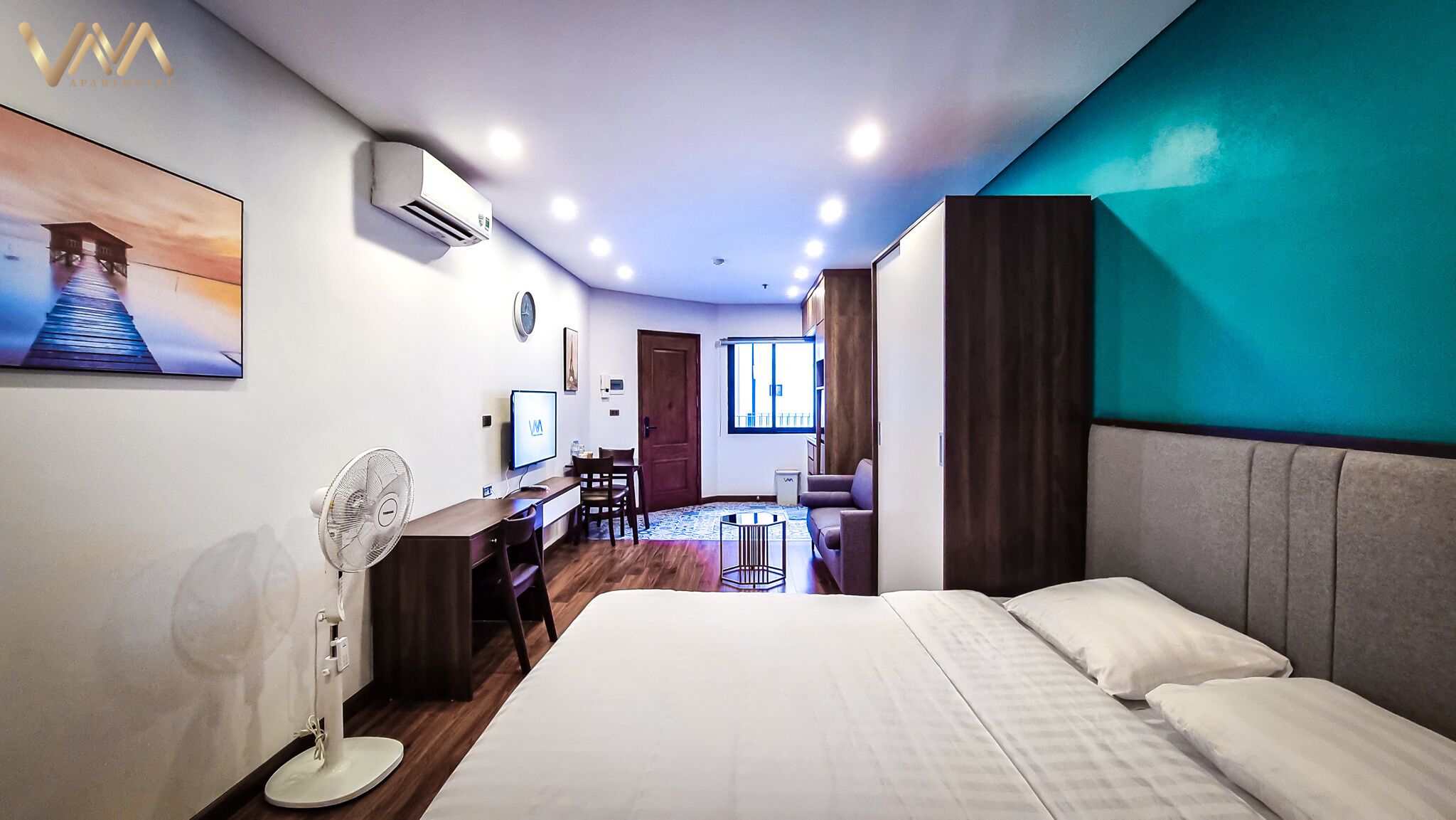 A hotel room with a bed, a desk, a chair, a TV, and an air conditioning unit.