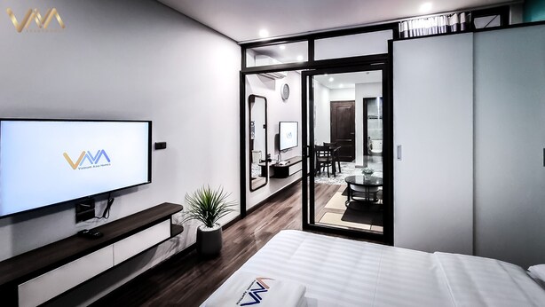 Una camera da letto moderna con una TV che mostra 'VVA Vietnam Asia Homes', un comodino, una pianta e un letto con un cuscino con logo.