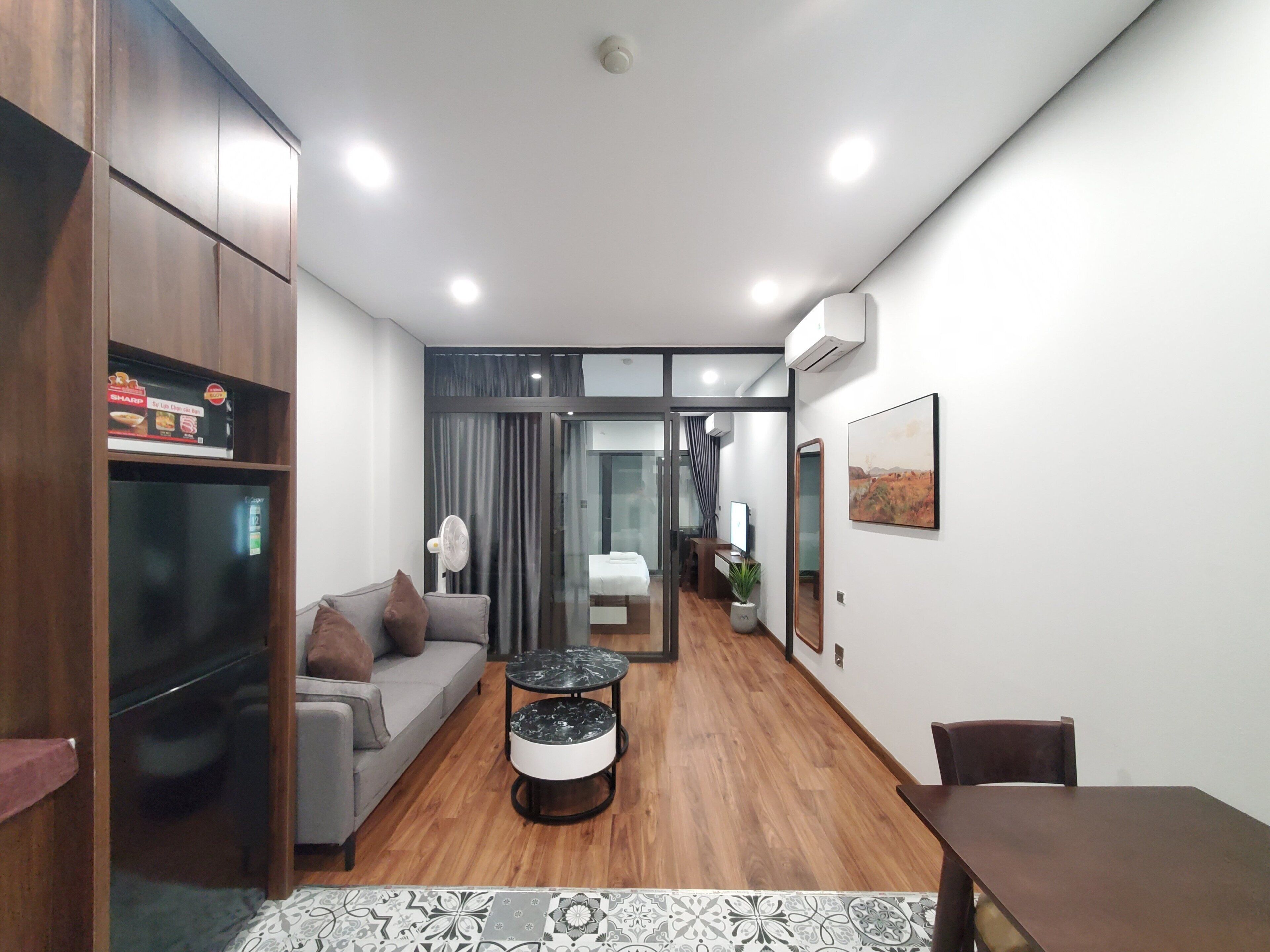 A modern hotel room with a sofa, a small round table, a dining table, a mirror, and a view into the adjacent bedroom.