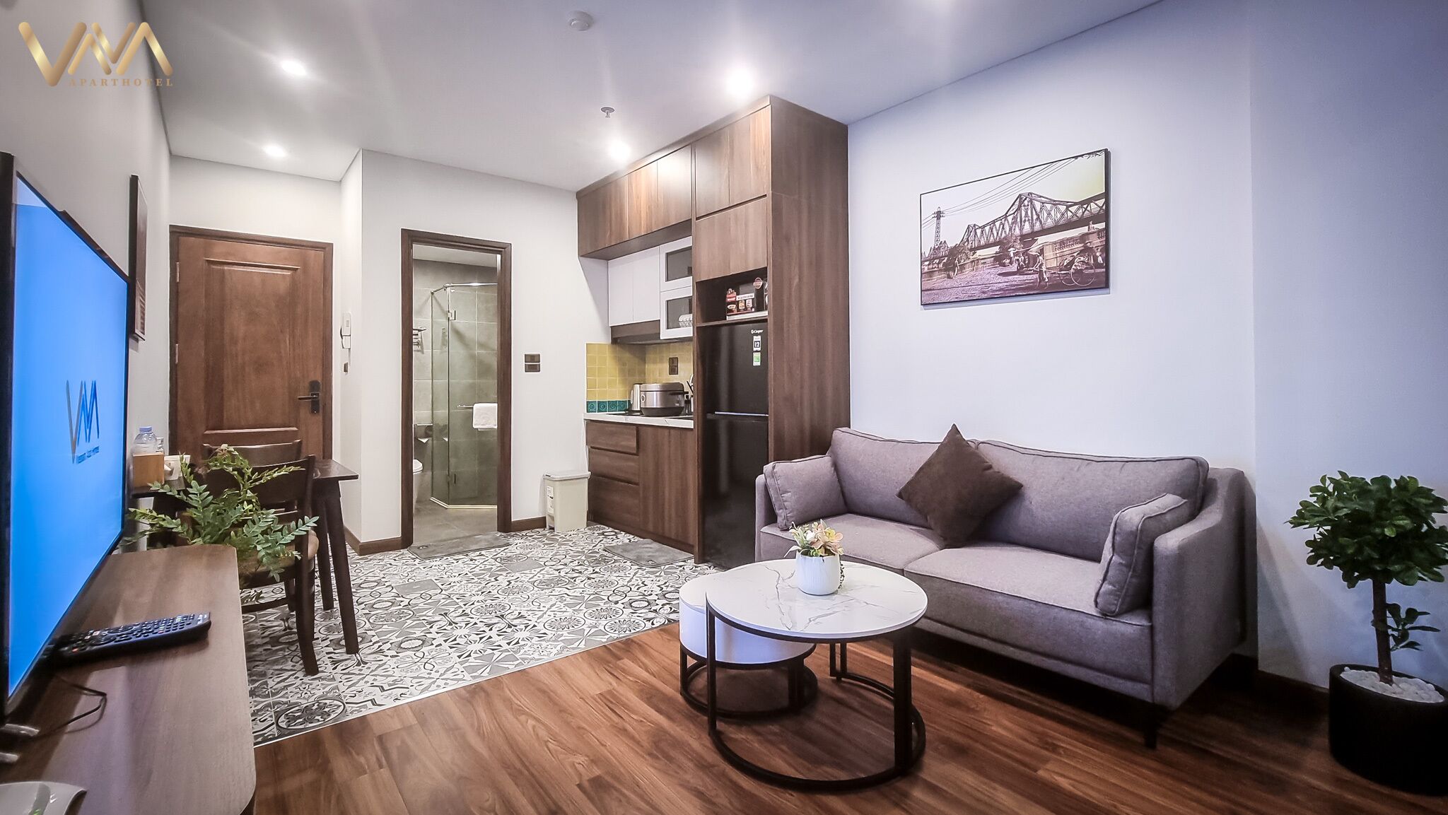 A modern living room with a sofa, coffee table, and a TV. There is a kitchen area with cabinets and a refrigerator. A bathroom with a glass shower door is visible.
