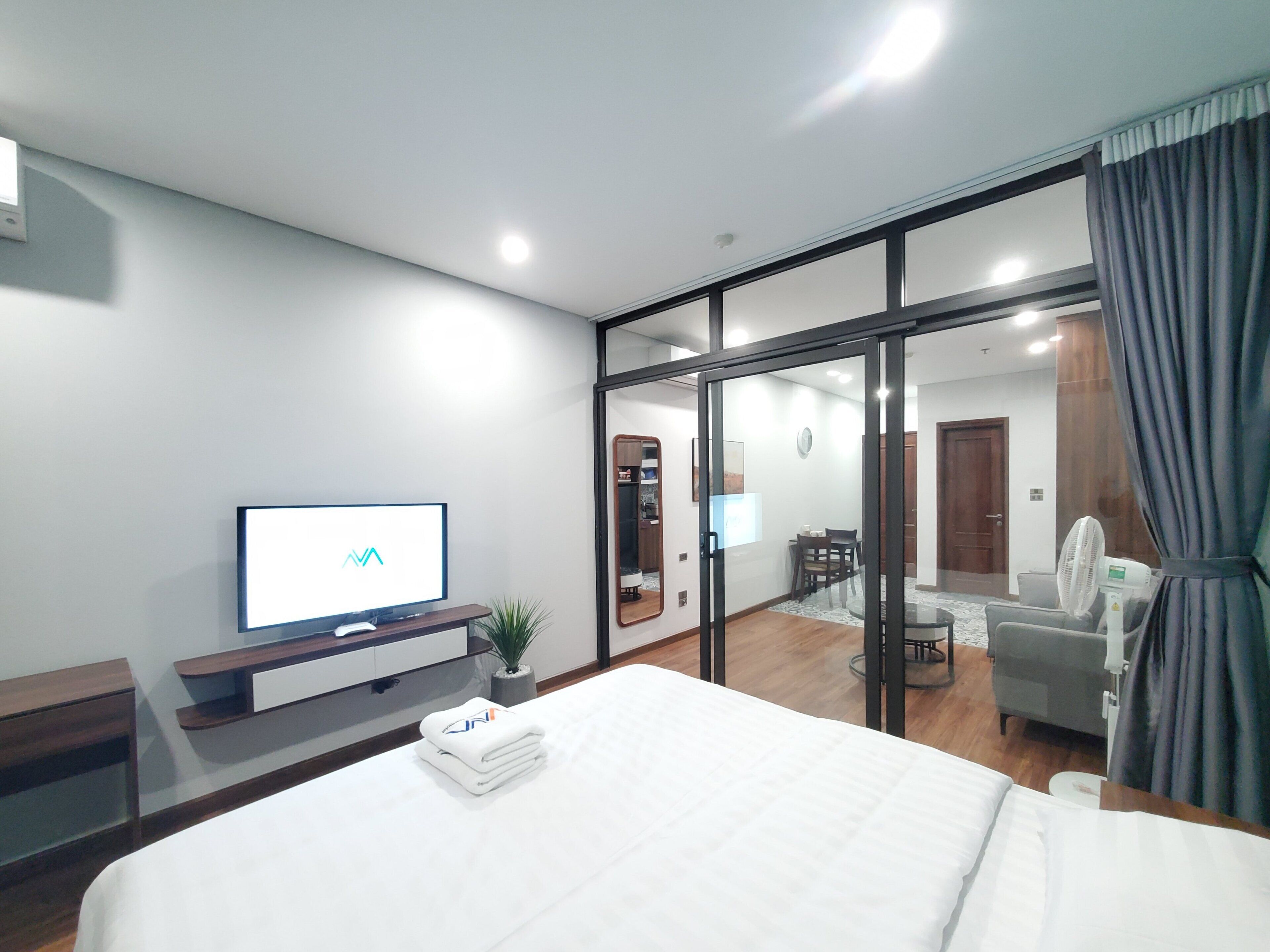 A modern hotel room with a large bed, a flat-screen TV, a desk with a plant, and a sliding glass door leading to a dining area.