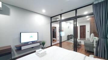 A modern hotel room with a large bed, a flat-screen TV, a desk with a plant, and a sliding glass door leading to a dining area.