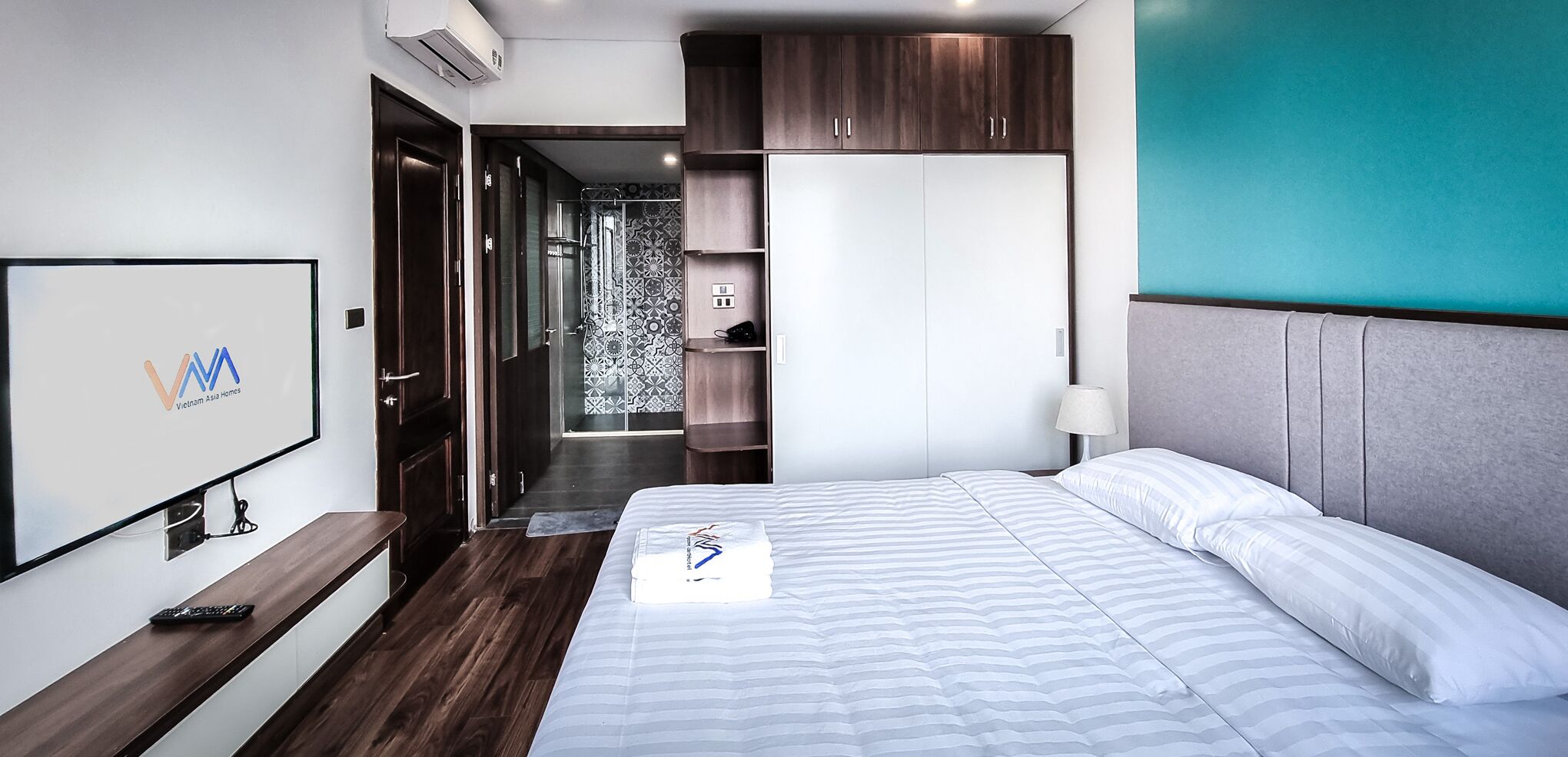 Ein modernes Hotelzimmer mit Bett, Fernseher, Kleiderschrank und einem durchgehenden Türbogen einsehbarer Badezimmer.