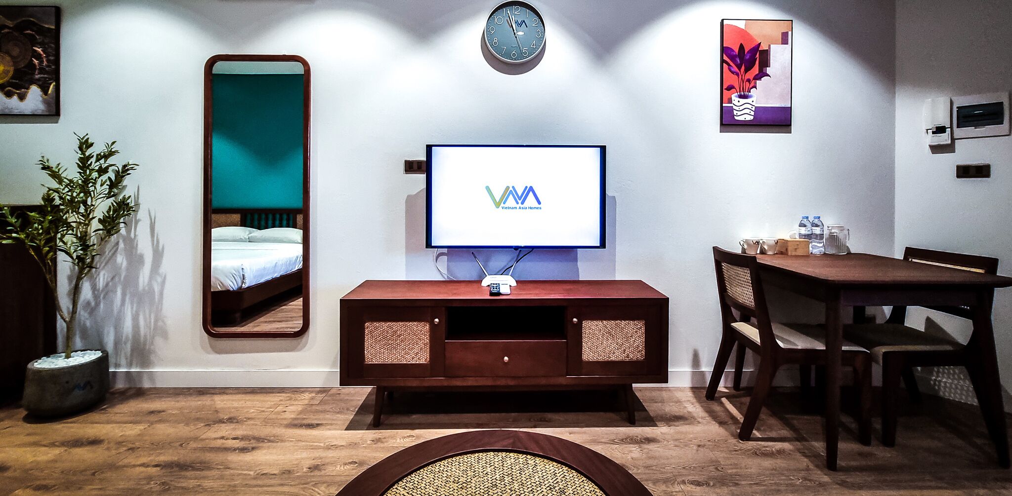 A hotel room with a bed, a TV, a clock, and a dining area with chairs and a table.