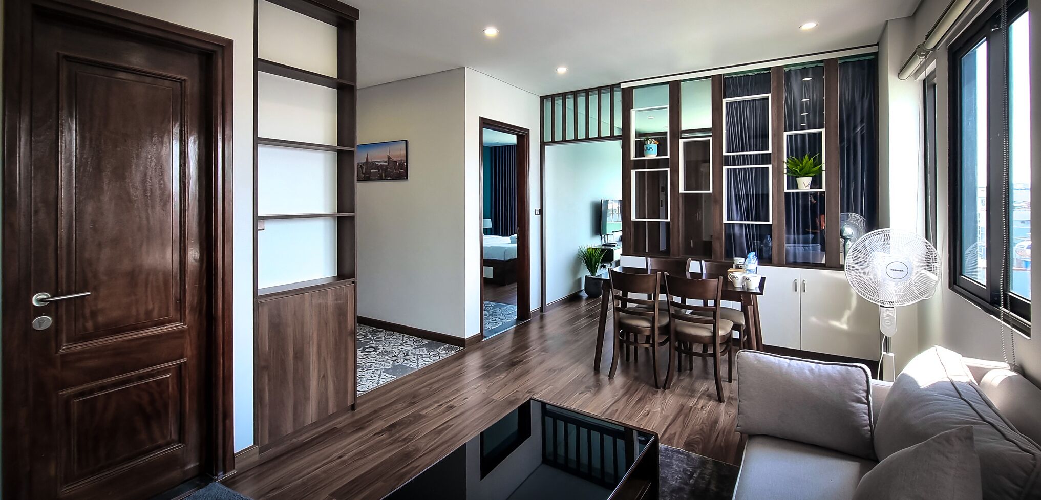 A modern living room with a wooden floor, a sofa, a dining table with chairs, a TV, and a large window with curtains.