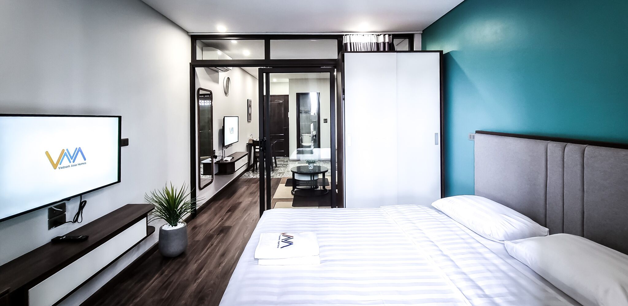 A modern hotel room with a bed, a TV, a desk, and a potted plant.