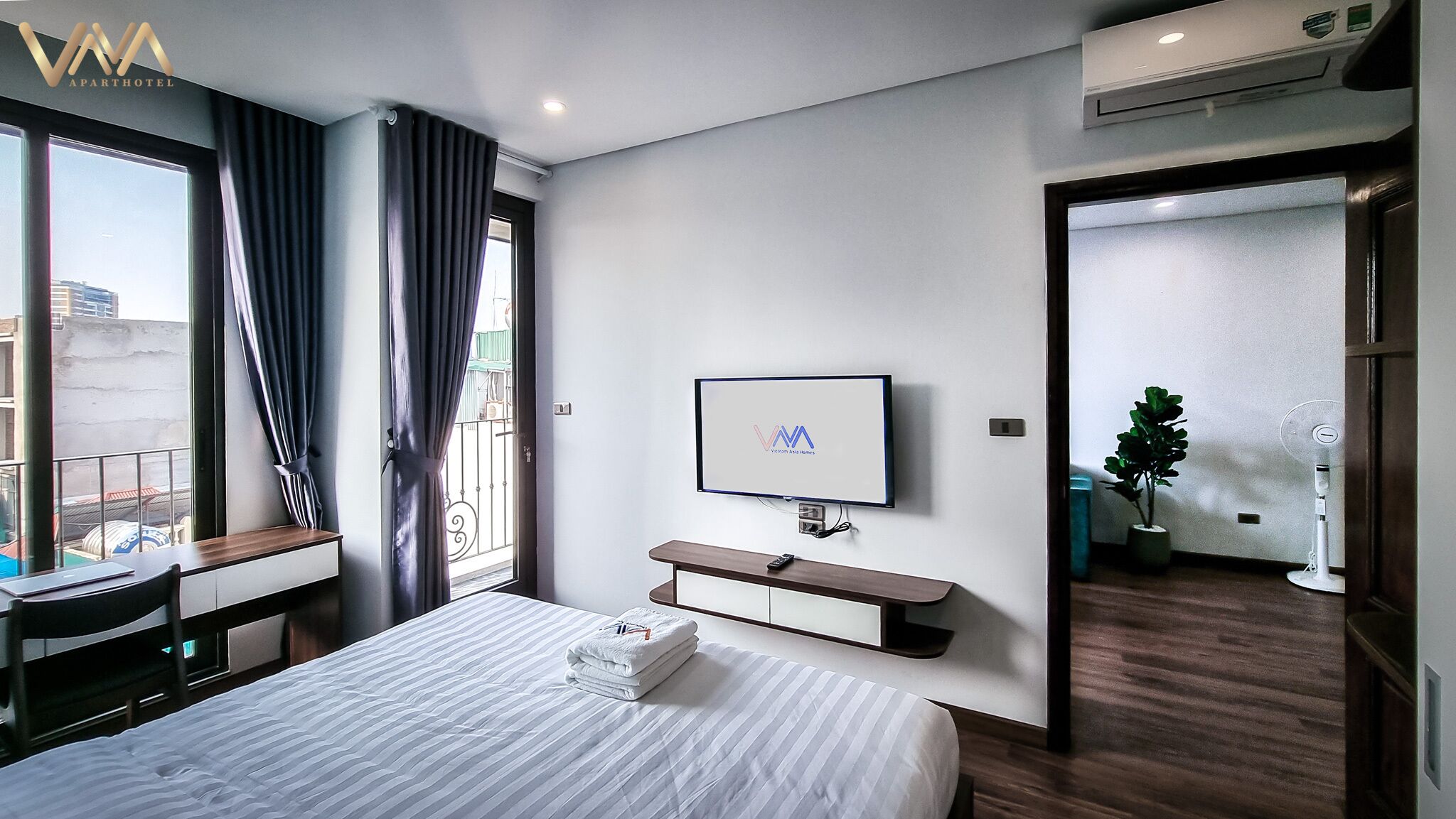 A modern hotel room with a large bed, a desk, a TV, and a balcony with curtains.