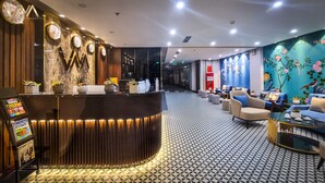 Un'area reception di un hotel dal design moderno, con una scrivania a reception dalle linee curve, diverse pendole e un'area salotto con divani e un tavolino.