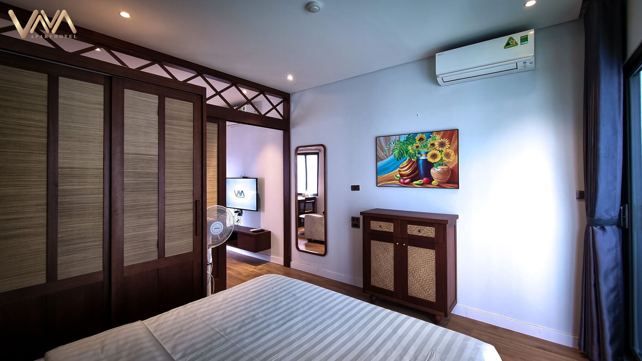 A bedroom with a bed, a TV, a mirror, a painting, and an air conditioning unit.