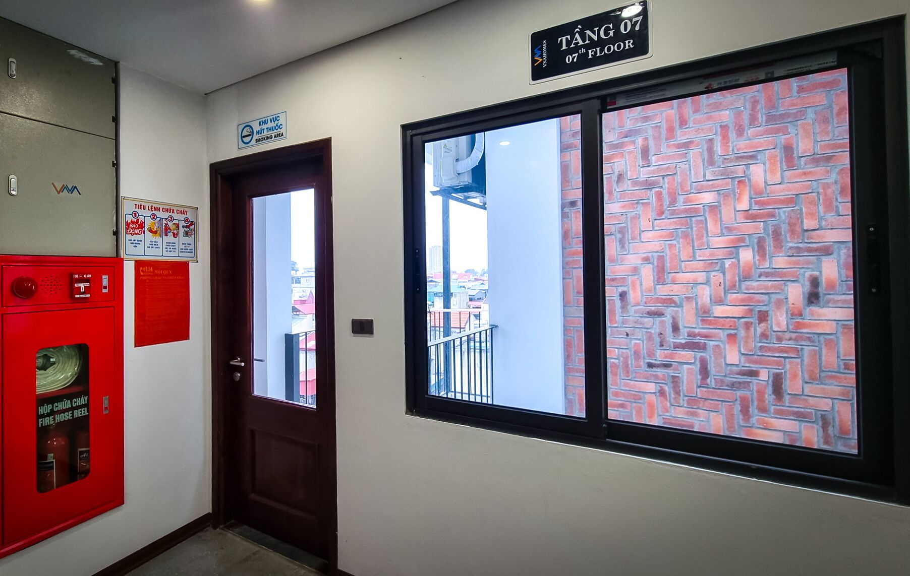 A hotel room with a door labeled "TANG 07 7th Floor," a fire hose reel cabinet, and a window with a brick pattern.