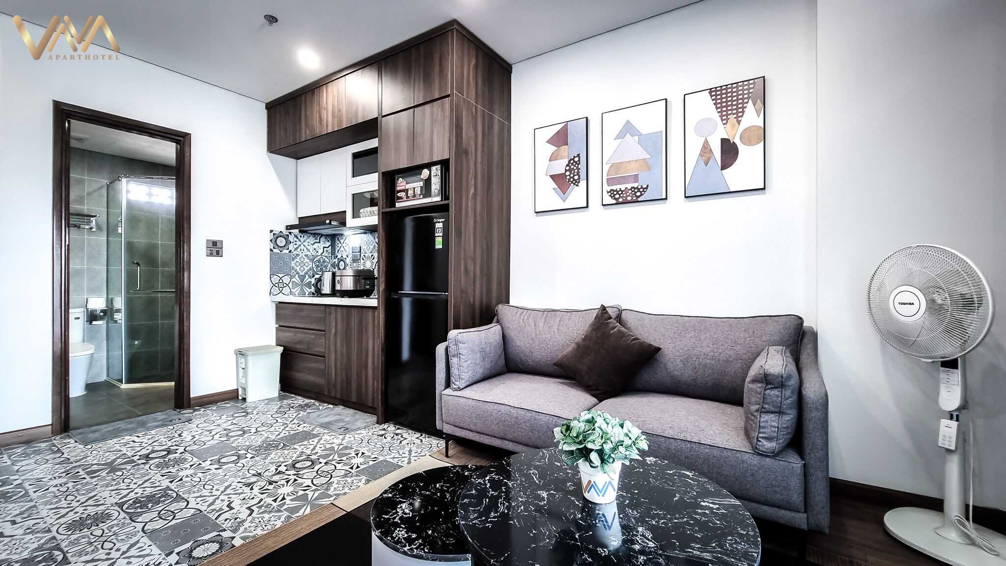 A modern living room with a grey sofa, a black coffee table with a vase, a Toshiba fan, and a kitchen area with a refrigerator and microwave.