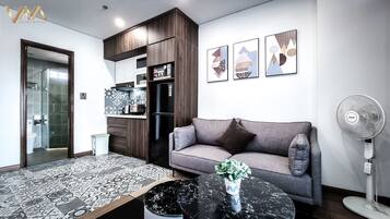 A modern living room with a grey sofa, a black coffee table with a vase, a Toshiba fan, and a kitchen area with a refrigerator and microwave.