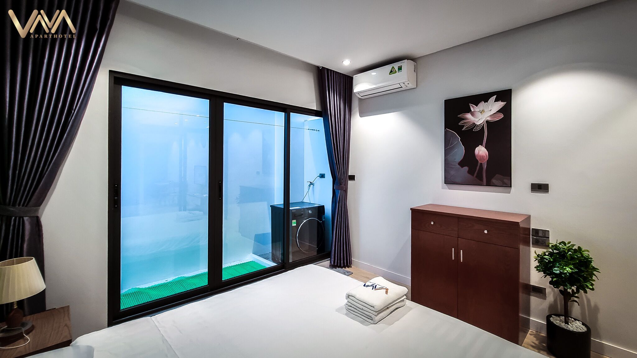 A modern bedroom with a large sliding glass door leading to a balcony, a bed with neatly folded towels, a dark wooden dresser, and a potted plant.