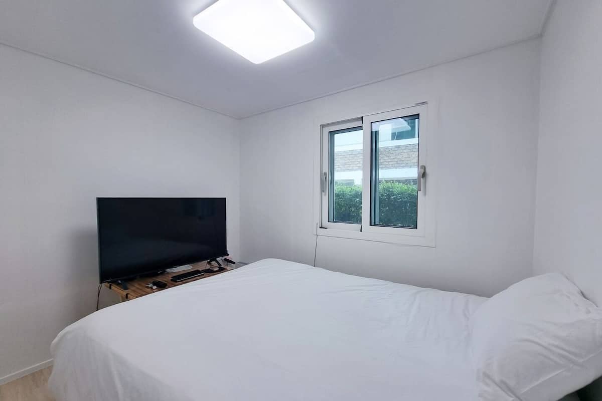 Een slaapkamer met een bed, een televisie op een standaard, een raam en een plafondlamp.