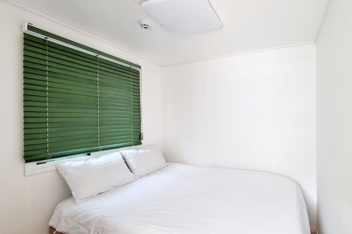 Een slaapkamer met een bed, witte muren en een raam met groene jaloezieën.