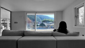 Uma sala de estar moderna com um sofá cinzento, uma porta de correr de vidro que dá para um jardim com árvores e um carro estacionado.