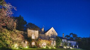 A large, historic building with multiple windows and a prominent central tower, surrounded by well-maintained gardens and outdoor seating areas.